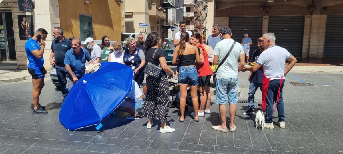 Donna sull’ asfalto in attesa dell’ambulanza riparata dal sole con un ombrello da un passante - 